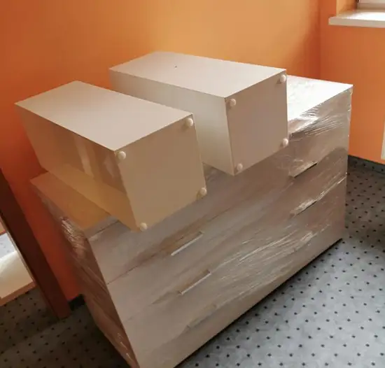 Zwei weiße, aufeinandergestapelte Aufbewahrungsboxen aus Holz, die auf einem sorgfältig mit Folie verpackten Möbelstück stehen, bereit für den Umzug